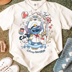 May include: Cream-colored t-shirt featuring a cartoon Stitch character in a captain's uniform, holding a compass. The shirt has the text "Let's Cruise 2025" and "Disney Adventure" on it. A Mickey Mouse balloon and nautical elements are also included in the design.