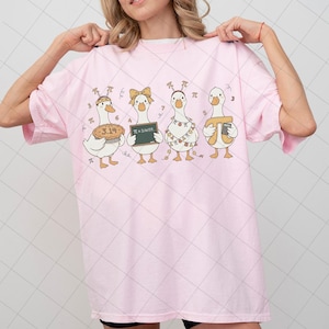 May include: Pink t-shirt featuring a whimsical design of four cartoon geese, each holding a different representation of pi. One goose holds a pie, another a chalkboard with "π = 3.14159", and others with pi symbols. A fun, educational graphic tee.