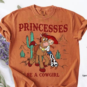 May include: Orange t-shirt with a graphic featuring a cartoon cowgirl hugging a horse, with a desert landscape backdrop. The text reads "PRINCESSES BE A COWGIRL" in a western-style font.