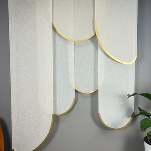 May include: White fabric window blinds with gold-colored curved accents. The blinds are hanging against a gray wall. The design features a layered, semi-circular pattern, adding a modern touch to the window treatment.