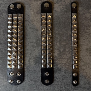 May include: Three black leather wristbands with silver pyramid studs. The wristbands vary in width and stud arrangement, with snap closures. The background is a gray, textured surface.