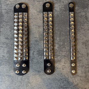 May include: Three black leather wristbands with silver pyramid studs. The wristbands vary in width and stud arrangement, fastened with silver snaps. The wristbands are displayed on a gray surface.