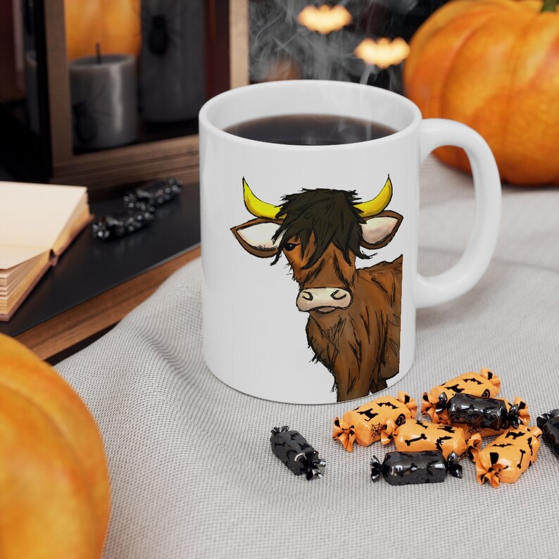 K&ouml;nnte beinhalten: Wei&szlig;e Keramiktasse mit einer Illustration einer braunen Highland-Kuh mit gelben H&ouml;rnern und schwarzen Haaren. Die Tasse ist mit dunkler Fl&uuml;ssigkeit gef&uuml;llt. Halloween-S&uuml;&szlig;igkeiten sind um die Tasse herum verstreut.