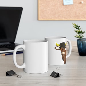 K&ouml;nnte beinhalten: Zwei wei&szlig;e Keramik-Kaffeetassen. Eine Tasse zeigt eine Cartoon-Illustration einer braunen Highland-Kuh mit gelben H&ouml;rnern und schwarzem Haar. Die andere Tasse ist schlicht wei&szlig;. Beide Tassen haben einen gebogenen Henkel.