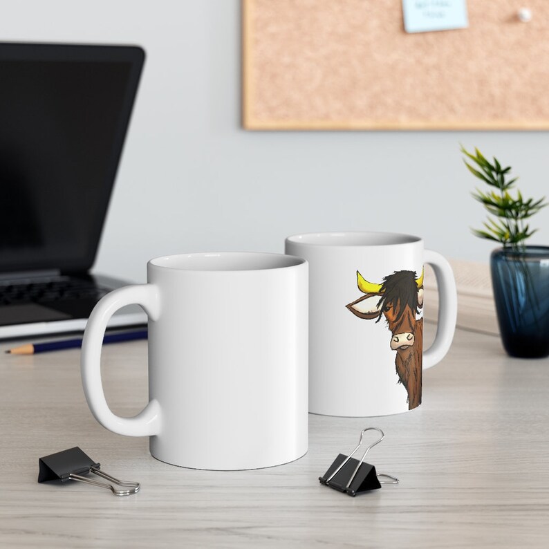 K&ouml;nnte beinhalten: Zwei wei&szlig;e Keramik-Kaffeetassen. Eine Tasse zeigt eine Cartoon-Illustration einer braunen Highland-Kuh mit gelben H&ouml;rnern und schwarzem Haar. Die andere Tasse ist schlicht wei&szlig;. Beide Tassen haben einen gebogenen Henkel.