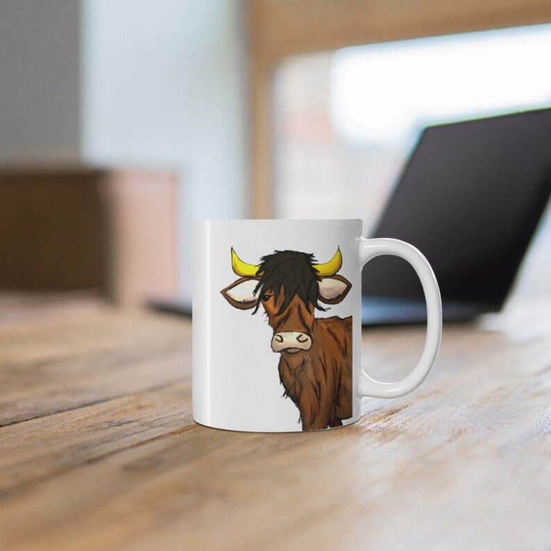 K&ouml;nnte beinhalten: Wei&szlig;e Keramiktasse mit einer Illustration einer Highland-Kuh. Die Kuh hat braunes Fell, gelbe H&ouml;rner und einen schwarzen Pony. Die Tasse hat einen gebogenen Henkel und steht auf einer Holzoberfl&auml;che.