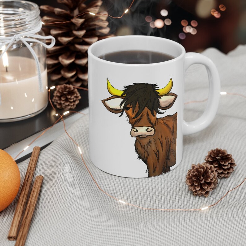 K&ouml;nnte beinhalten: Wei&szlig;e Keramik-Kaffeetasse mit einer Cartoon-Highland-Kuh. Die Kuh hat braunes Fell, gelbe H&ouml;rner und schwarzes Haar. Die Tasse ist mit einer dunklen Fl&uuml;ssigkeit gef&uuml;llt und hat einen gebogenen Henkel.