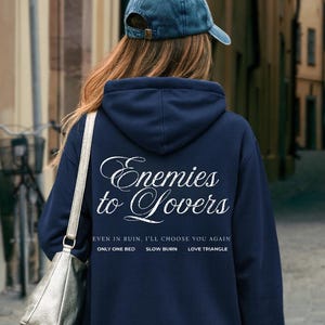 May include: Navy blue hoodie with the text "Enemies to Lovers" in white script. Below the main text are the phrases "EVEN IN RUIN, I'LL CHOOSE YOU AGAIN", "ONLY ONE BED", "SLOW BURN", and "LOVE TRIANGLE". The person is wearing a blue baseball cap.