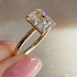 Puede incluir: Un anillo de oro con un diamante de talla radiante. El diamante de forma cuadrada está sujeto por cuatro garras. El anillo está sostenido por una mano. El anillo es una joya.