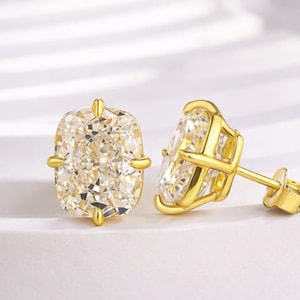 May include: A pair of gold-colored stud earrings with large, cushion-cut, light-yellow gemstones. The earrings are set in a four-prong setting with a post and butterfly backing. The gemstones have a brilliant cut.