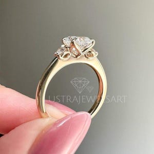 Puede incluir: Un anillo de oro con tres piedras preciosas transparentes. El anillo lo sostiene una persona con esmalte de uñas rosa. El texto "LUSTRAJEWELSART" es visible en el anillo.