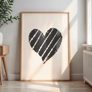 May include: A framed art print featuring a large black heart with white diagonal stripes on a cream background. The artwork is in a natural wood frame and is displayed on a wooden floor.