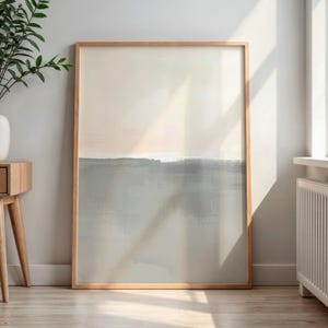 May include: Abstract art print in a natural wood frame. The artwork features a soft colour palette with a light beige upper section and a grey-blue lower section, separated by a horizontal line. The painting has a minimalist style, suitable for home decor.