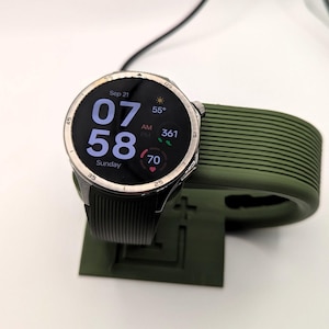 May include: A silver smartwatch with a black strap shows the time as 07:58 on Sunday, September 21st. The watch is on a green charging stand. The screen displays the temperature and other health metrics.