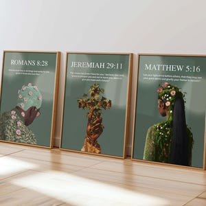 May include: Three framed art prints with gold frames. The prints feature illustrations of people with floral and plant elements, set against a muted green background. Each print has a Bible verse and title. The prints are displayed on a wooden floor.