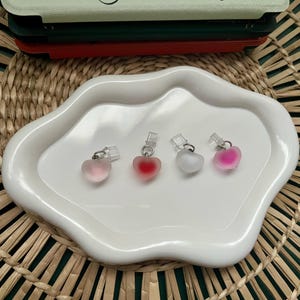 Op de afbeelding: Vier hartvormige hangers in roze en rode tinten, gepresenteerd op een wit dienblad met golvende randen. Elke hanger heeft een kleine zilverkleurige beugel. De hangers zijn gerangschikt op een geweven, natuurlijk getinte ondergrond.