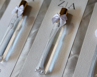 Op de afbeelding: Doorzichtige glazen parfumflesjes met gouden doppen en witte strikjes zijn gerangschikt op lichtgrijze kaarten. De kaarten hebben de naam "ZIA" bovenaan en de namen "Zeynep & Ali" met de datum "11.10.2025" onderaan.
