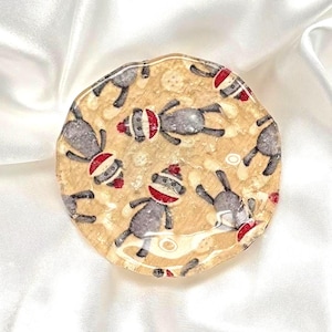 May include: A round coaster with a beige background featuring a pattern of gray sock monkey figures wearing red hats. The coaster has a clear, glossy finish and an irregular edge.