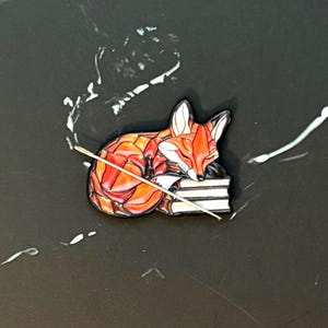 Könnte beinhalten: Ein farbenfroher Emaille-Pin mit einem schlafenden Fuchs, der auf einem Stapel Bücher ruht. Der Fuchs ist hauptsächlich orange und rot mit weißen Akzenten. Der Pin hat eine schwarze Umrandung und ein kleiner Stock liegt auf den Büchern.