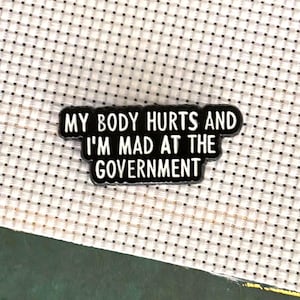 Könnte beinhalten: Ein schwarzer Emaille-Pin mit weißem Text, der lautet: "MY BODY HURTS AND I'M MAD AT THE GOVERNMENT." Der Pin ist rechteckig und vor einem weiß-schwarz gepunkteten Hintergrund platziert.