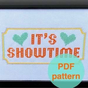 May include: Cross-stitch pattern framed in black. The design features the words "IT'S SHOWTIME" in orange, with green heart accents. The text is on a light blue background. A teal circle with the words "PDF pattern" is in the lower right corner.