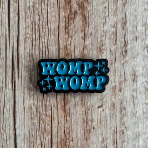 Può includere: Un ciondolo per scarpe nero e blu con la parola "WOMP" in un design impilato. La parola è di colore blu brillante con accenti di stelle. Il ciondolo è su uno sfondo di legno.