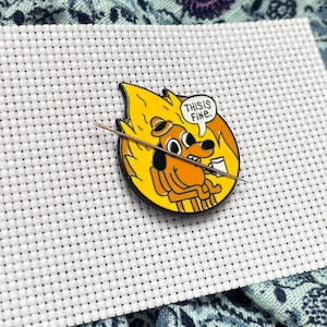 Könnte beinhalten: Ein runder Emaille-Pin mit einem Cartoon-Hund, der in einem brennenden Raum sitzt, mit dem Text "THIS IS FINE" in einer Sprechblase. Der Pin ist gelb, orange und schwarz, mit einer silbernen Nadel.