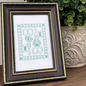 May include: A framed cross-stitch featuring green sewing tools: scissors, thread spools, and a needle. The frame is dark brown with a gold inner edge. A potted plant with green leaves sits in the background on a wooden surface.