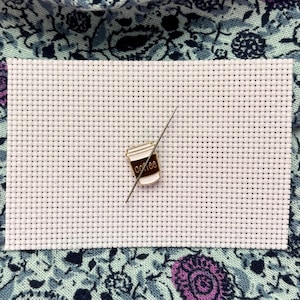 Könnte beinhalten: Ein weißer Kreuzstichstoff mit einer Kaffeetassen-Anstecknadel und einer Nadel. Die Anstecknadel ist eine Miniatur-Kaffeetasse mit dem Wort "coffee" darauf. Die Nadel ist silberfarben und wird durch den Stoff gesteckt.