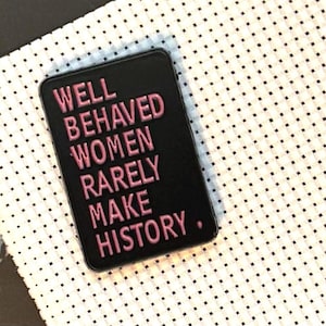Könnte beinhalten: Ein schwarzer rechteckiger Anstecker mit dem pinkfarbenen Schriftzug "WELL BEHAVED WOMEN RARELY MAKE HISTORY". Der Anstecker liegt auf einem weißen Stoff mit schwarzen Punkten und einer schwarzen Oberfläche.