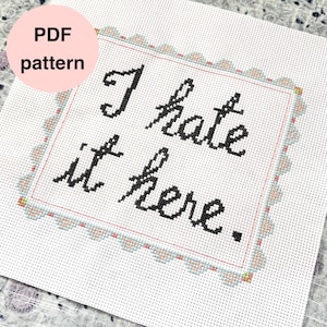 May include: A cross-stitch design featuring the phrase "I hate it here" in black script. The design is framed with a pink border and a decorative edge. A pink circle with "PDF pattern" is in the upper left corner.