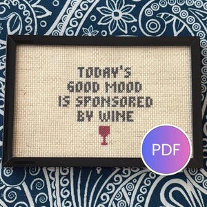 Puede incluir: Un bordado de punto de cruz rectangular en un marco negro con el texto "TODAY'S GOOD MOOD IS SPONSORED BY WINE" en hilo negro, acompañado de un pequeño icono de copa de vino rojo bordado bajo el texto sobre un fondo de tela beige claro.