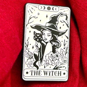 Könnte beinhalten: Rechteckiger Anstecker mit einer schwarz-weißen Illustration einer Hexe, die eine schwarze Katze hält. Die Hexe trägt einen Hut und ist von floralen Elementen umgeben. Der Text "THE WYTCH" steht unten.