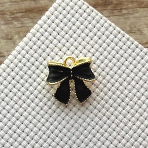 Könnte beinhalten: Ein kleiner, schwarzer Schleifenanhänger mit goldenen Rändern. Der Anhänger liegt auf einem weißen, gewebten Hintergrund. Der Anhänger hat eine Schlaufe zum Befestigen. Das Design der Schleife ist schlicht und elegant.