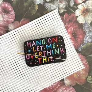 Könnte beinhalten: Eine schwarze rechteckige Anstecknadel mit dem Text "HANG ON. LET ME OVERTHINK THIS..." in bunten Buchstaben. Eine goldene Nadel ist in die Anstecknadel gesteckt. Die Anstecknadel befindet sich auf einem weißen Kreuzstichstoff.