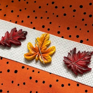 May include: Three autumn-themed leaf-shaped embellishments in red, orange, and yellow. One leaf has a needle through it. The leaves are on white cross-stitch fabric, set against an orange polka-dot background.