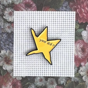 May include: A yellow star-shaped enamel pin with the text "your did it" in black lettering. The pin is set on a white cross-stitch fabric, with a floral background.