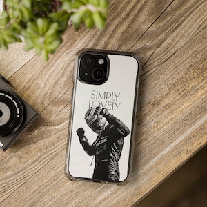 May include: Clear phone case featuring a black and white image of a racing driver with the text "SIMPLY LOVELY". The case is on a wooden surface, with a camera and plant visible in the background.