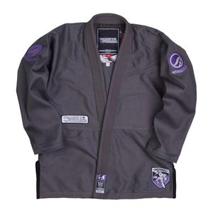 Pode incluir: Um gi cinzento de artes marciais com gola e acabamentos cinza escuro. O gi apresenta vários patches com texto e logotipos, incluindo "TRAINERS & FORMER" e "SHOYOROLL". O gi tem um ajuste sob medida e é projetado para treinamento e competição.
