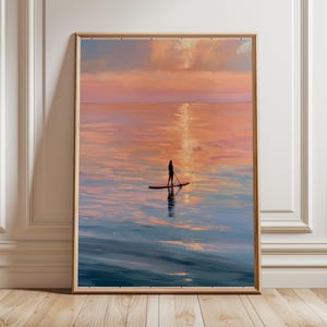 Op de afbeelding: Een ingelijste verticale wandkunstprint met het silhouet van een persoon op een paddleboard in het midden van een kalme oceaan bij zonsondergang. De lucht en het water vertonen zachte kleurovergangen van oranje, roze en blauw.