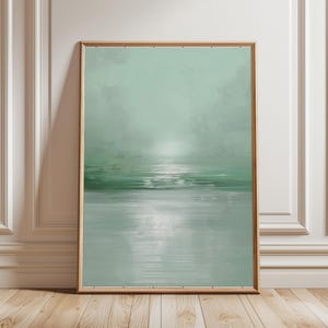 May include: Framed abstract landscape print. The artwork depicts a tranquil seascape in shades of green and grey, creating a calming atmosphere. The water reflects the sky, resulting in a minimalist aesthetic. Suitable for home decor, the print is in a wooden frame.