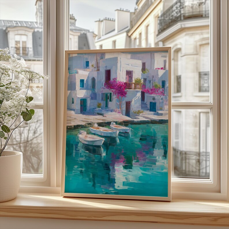 May include: A framed art print depicts a coastal scene with white buildings, blue doors and windows, and vibrant pink flowers. Three small boats float on the turquoise water. The artwork is displayed on a wooden windowsill.