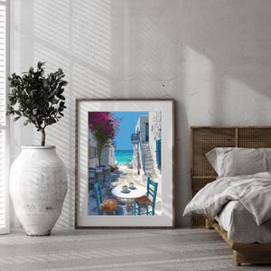 May include: A framed print depicts a coastal scene with white buildings, blue doors, and a turquoise sea. A table and chairs are set up on a cobblestone street, with pink flowers overhead. The artwork is displayed in a room with a large vase and a bed.