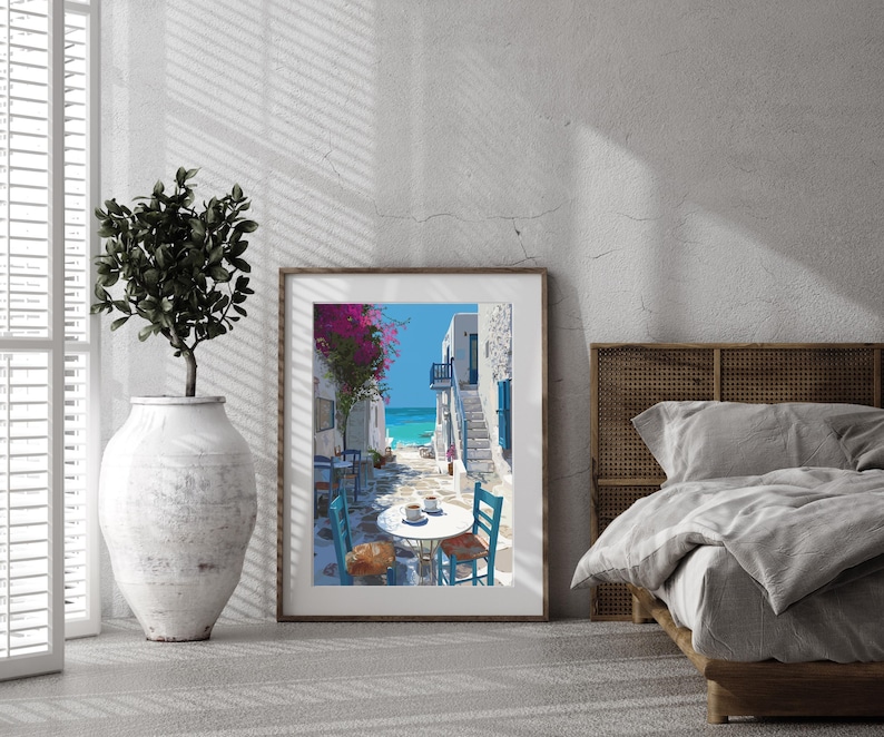 May include: A framed print depicts a coastal scene with white buildings, blue doors, and a turquoise sea. A table and chairs are set up on a cobblestone street, with pink flowers overhead. The artwork is displayed in a room with a large vase and a bed.