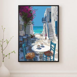 May include: A framed art print depicts a sunlit alleyway in a coastal town. White buildings with blue accents line the cobblestone street. A table with two cups of coffee sits in the foreground, with blue chairs. Pink flowers and the turquoise sea are visible.