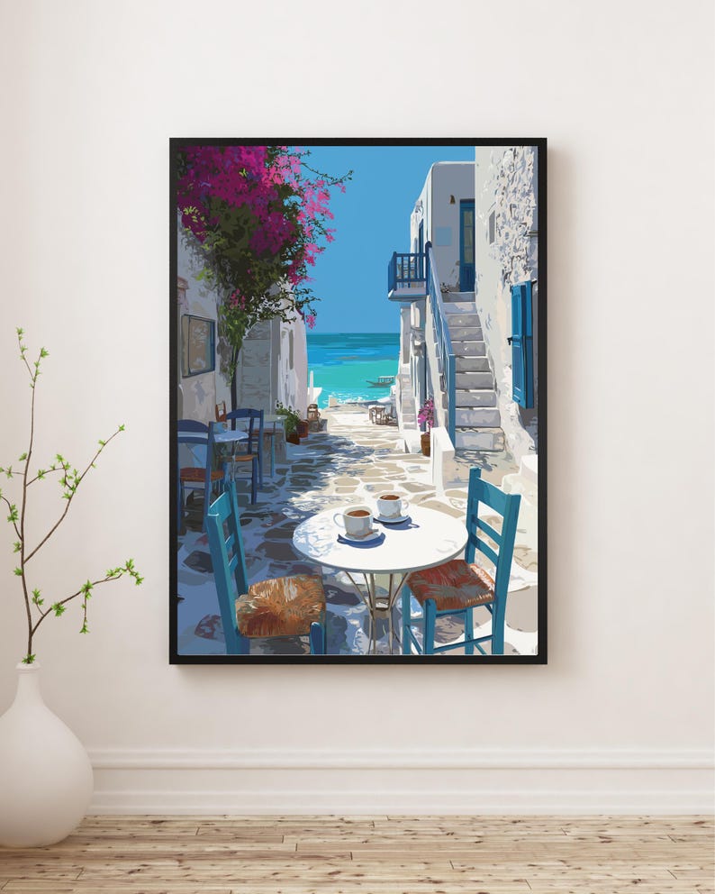 May include: A framed art print depicts a sunlit alleyway in a coastal town. White buildings with blue accents line the cobblestone street. A table with two cups of coffee sits in the foreground, with blue chairs. Pink flowers and the turquoise sea are visible.