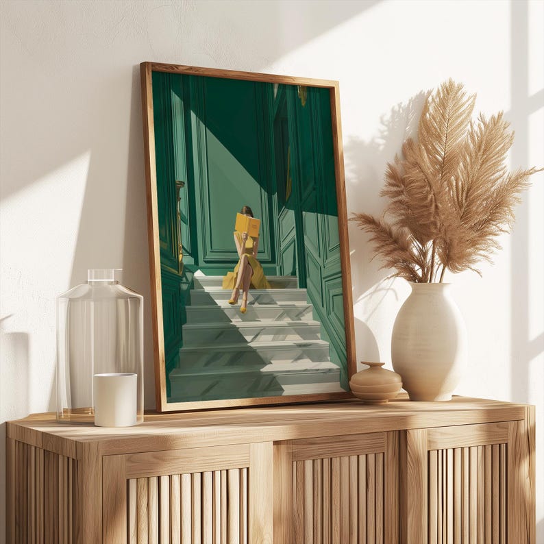 May include: A framed art print depicts a person in a yellow dress reading a book on a staircase. The artwork features a green and white color scheme with a wooden frame. A vase with dried plants sits on a wooden cabinet.