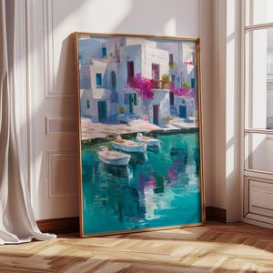 May include: A framed painting depicts a coastal scene with white buildings, turquoise water, and small boats. The artwork features a gold frame and is leaning against a white wall. Pink flowers and reflections add color to the serene landscape.