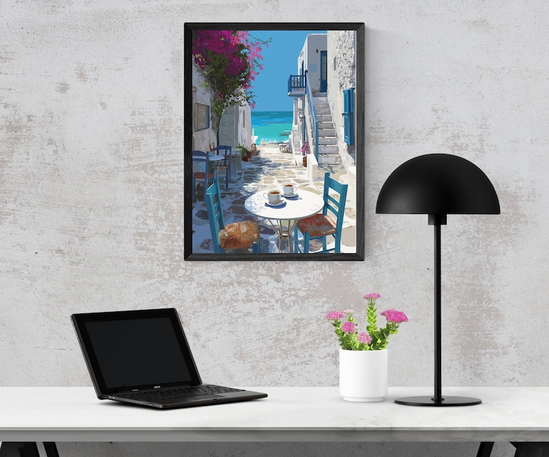 May include: A framed art print depicts a coastal scene with white buildings, blue shutters, and a turquoise sea. A table with two cups of coffee and blue chairs is in the foreground. A black desk lamp, a laptop, and a small white flower pot are also visible.
