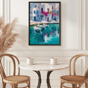 May include: A framed art print depicts a coastal scene with white buildings, pink flowers, and small boats on turquoise water. The artwork is displayed above a marble table with two white mugs and wicker chairs.